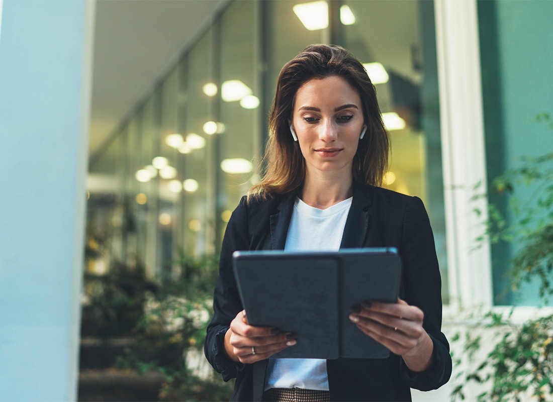 Blog - Portrait of a Young Business Woman Standing Outside a Modern Office Building While Looking at a Tablet She is Holding in Her Hands