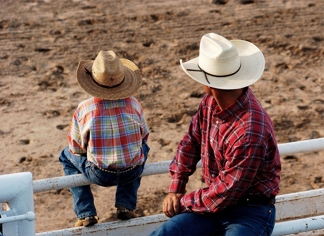 We Are Independent - Rear View of a Father and Son Wearing Cowboy Hats Sitting on a White Fence on a Ranch Looking Out at a Dirt Field During a Rodeo