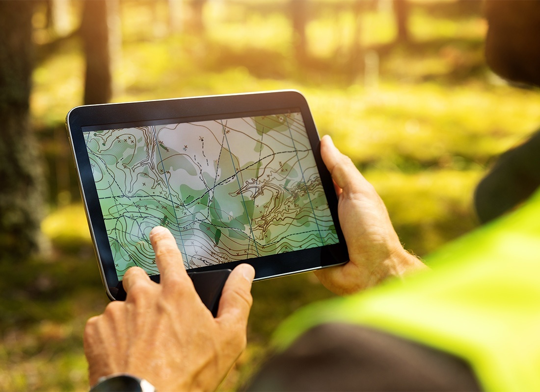 Surveyor Insurance - Closeup View of a Land Surveyor Looking at a Land Chart on a Tablet While Standing Outside on a Lot with Trees
